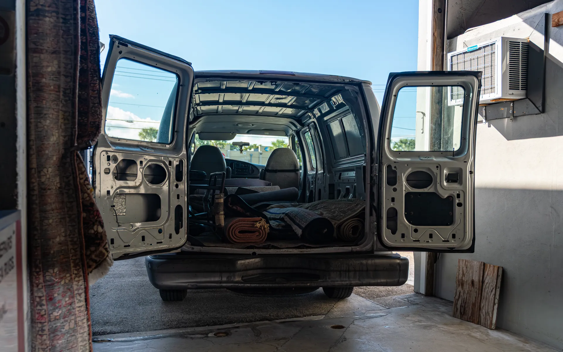 Several rolled rugs and a hand truck in the back of a white cargo van with its rear doors open at a shop bay