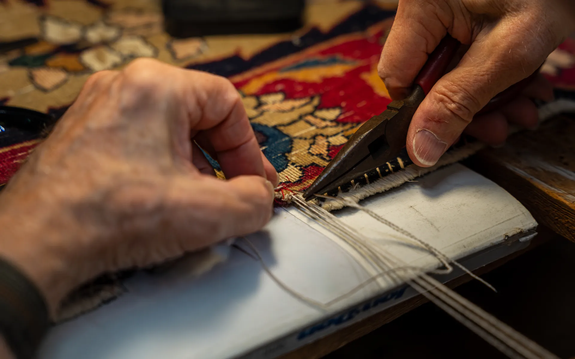 Hands use needle-nose pliers to pull a needle and white threads through the edge of a red and gold rug