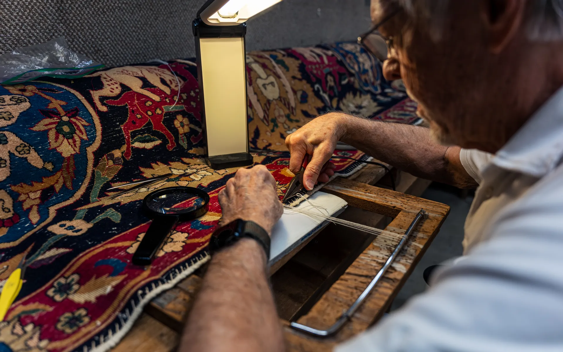 Rug repairer working on the edge of an oriental pictorial rug with pliers, a magnifier and a lamp at a workbench