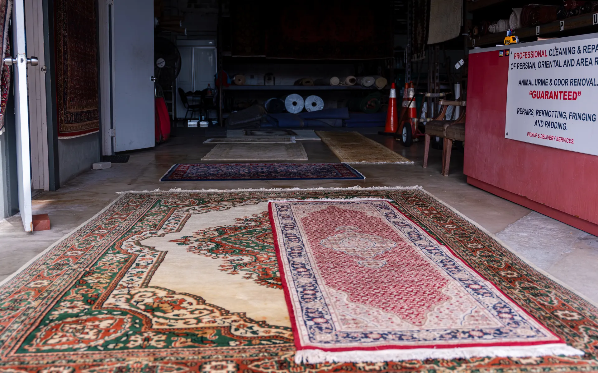 Oriental and area rugs laid flat on a rug shop floor beside a counter sign for cleaning, repair and pickup and delivery