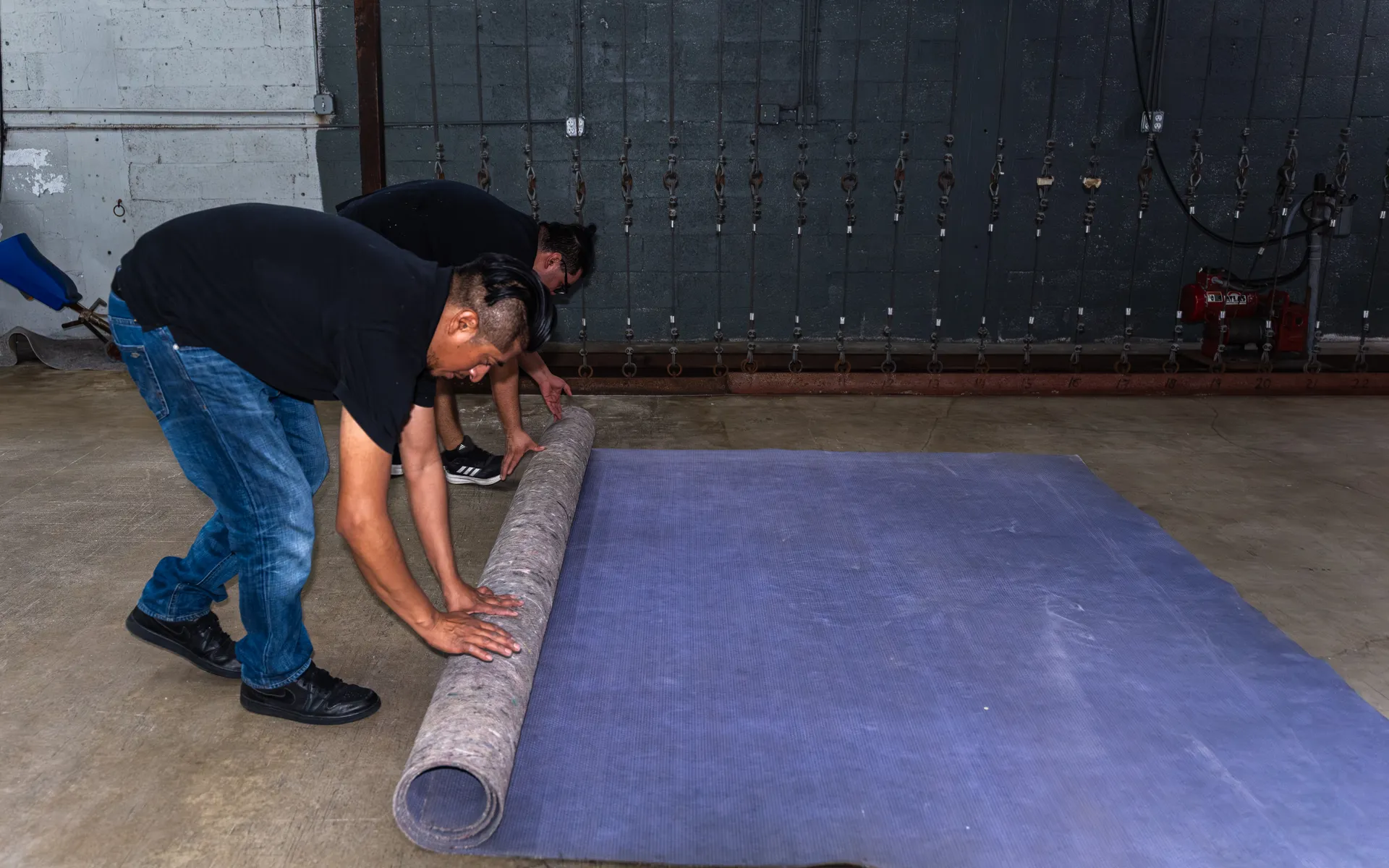 Two workers roll up a gray felt rug pad with a blue ribbed side on a concrete shop floor in front of hanging chains