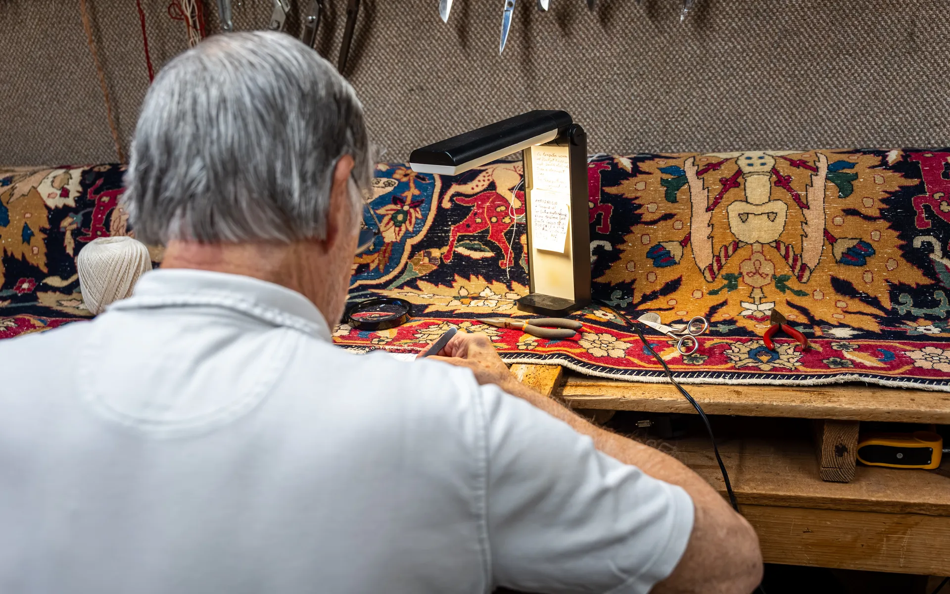 Person repairing a colorful antique-style rug by hand at a wooden workbench with a lamp, magnifier and scissors