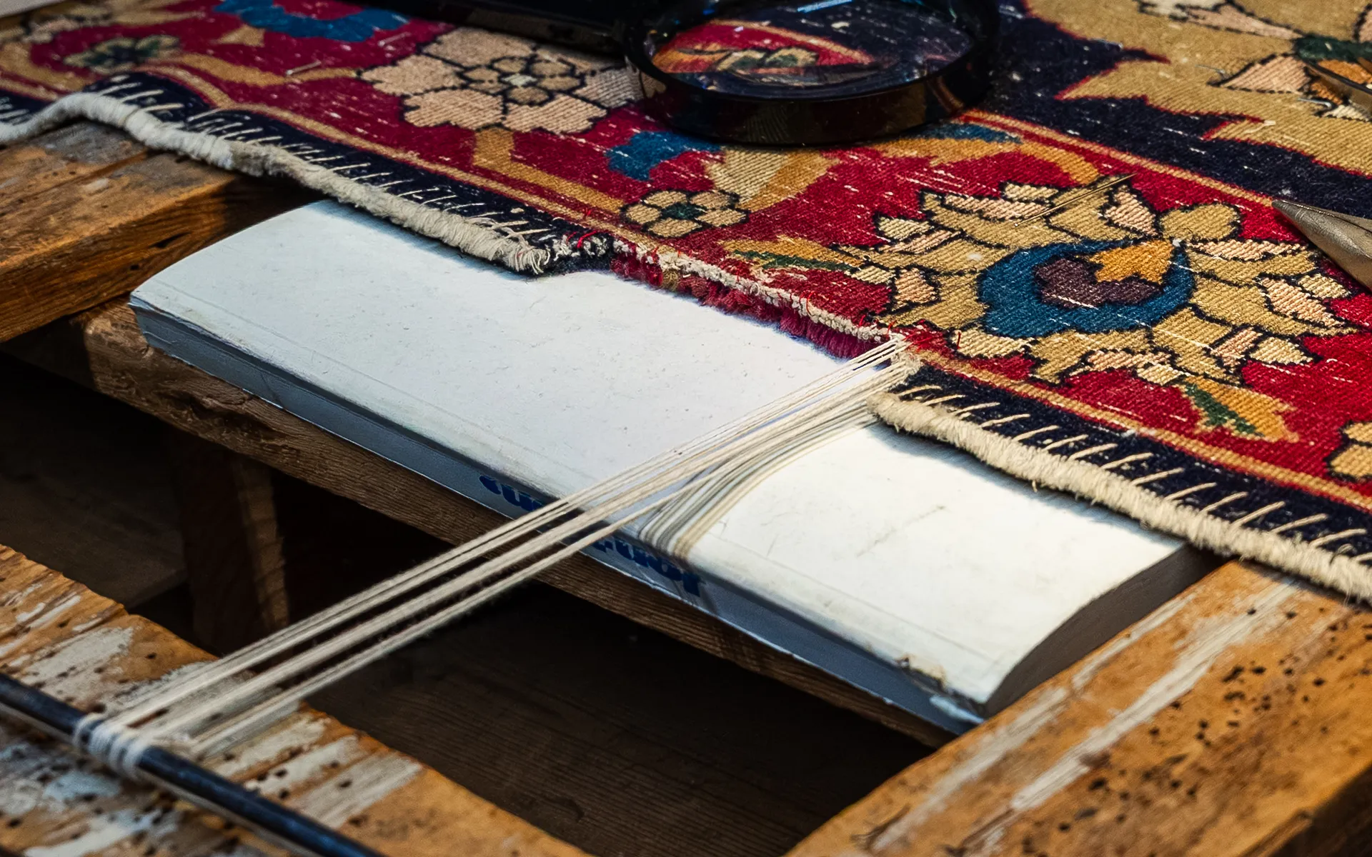 Close-up of a worn red and navy Persian-style rug edge under repair, with new cream warp threads strung to a metal rod