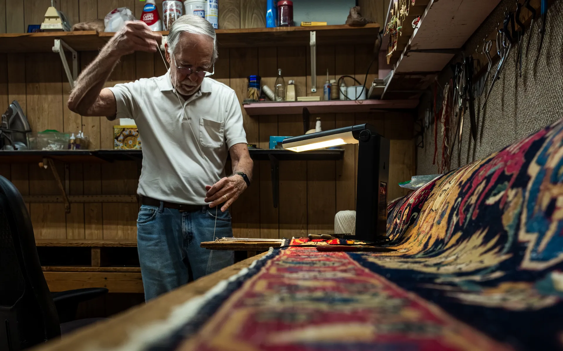 Craftsman in a white polo pulling a thread taut while repairing a large red and navy Persian-style rug at a workbench